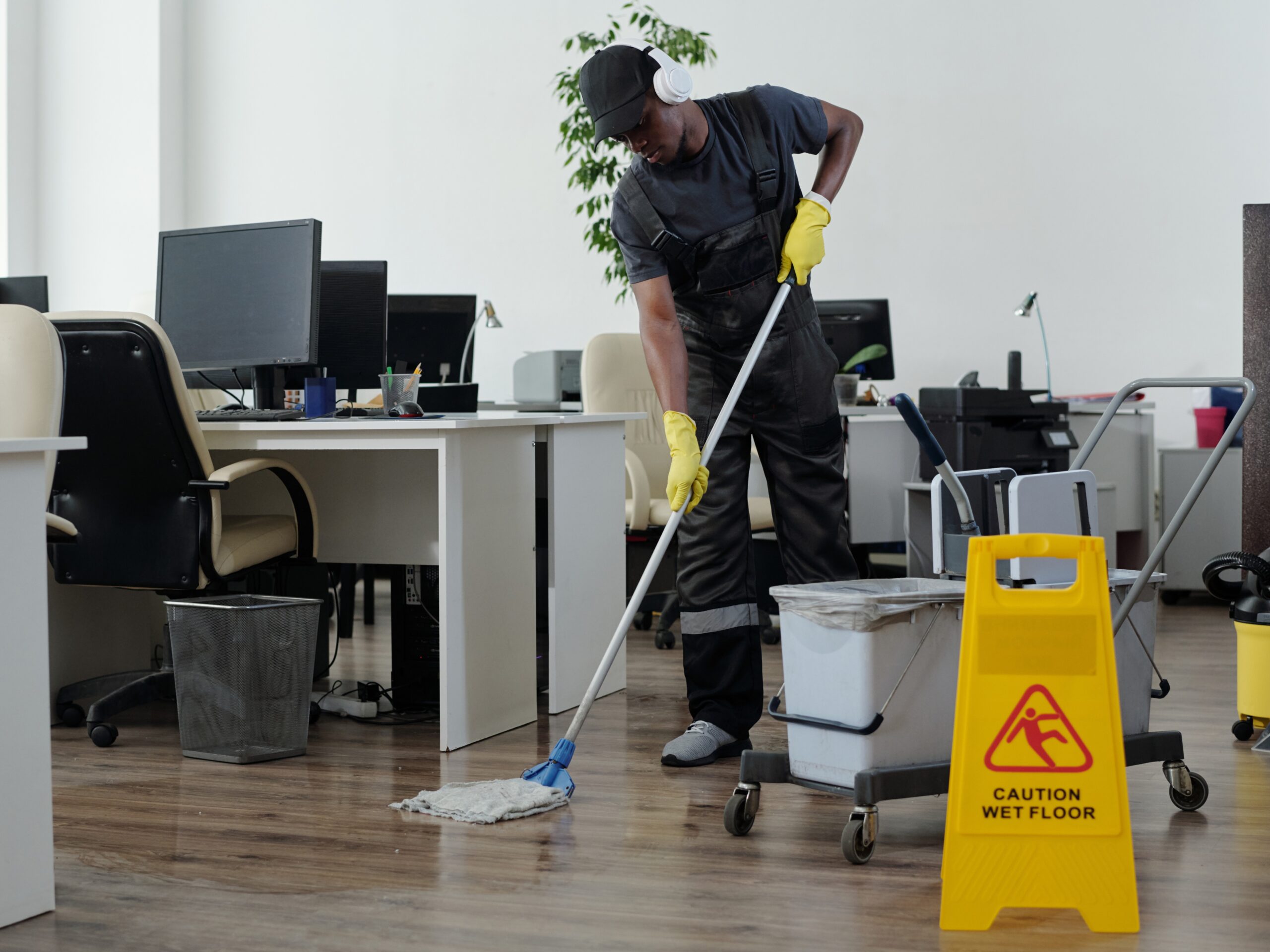 Contemporary young black man in workwear cleaning floor in openspace office in front of yellow plastic signboard with caution
