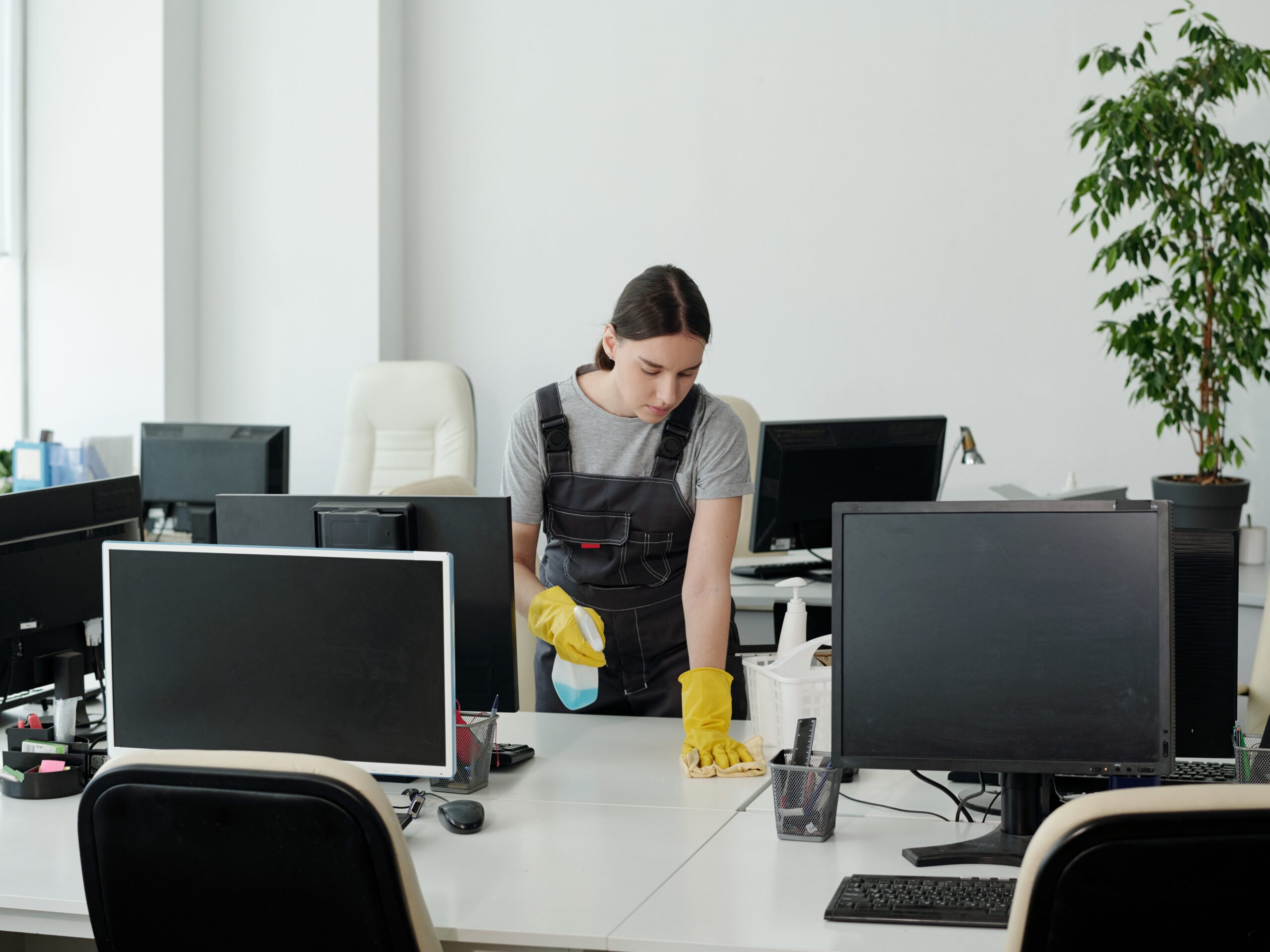 Young woman in coveralls and gloves using detergent while wiping one of desks with computer monitors in large openspace office