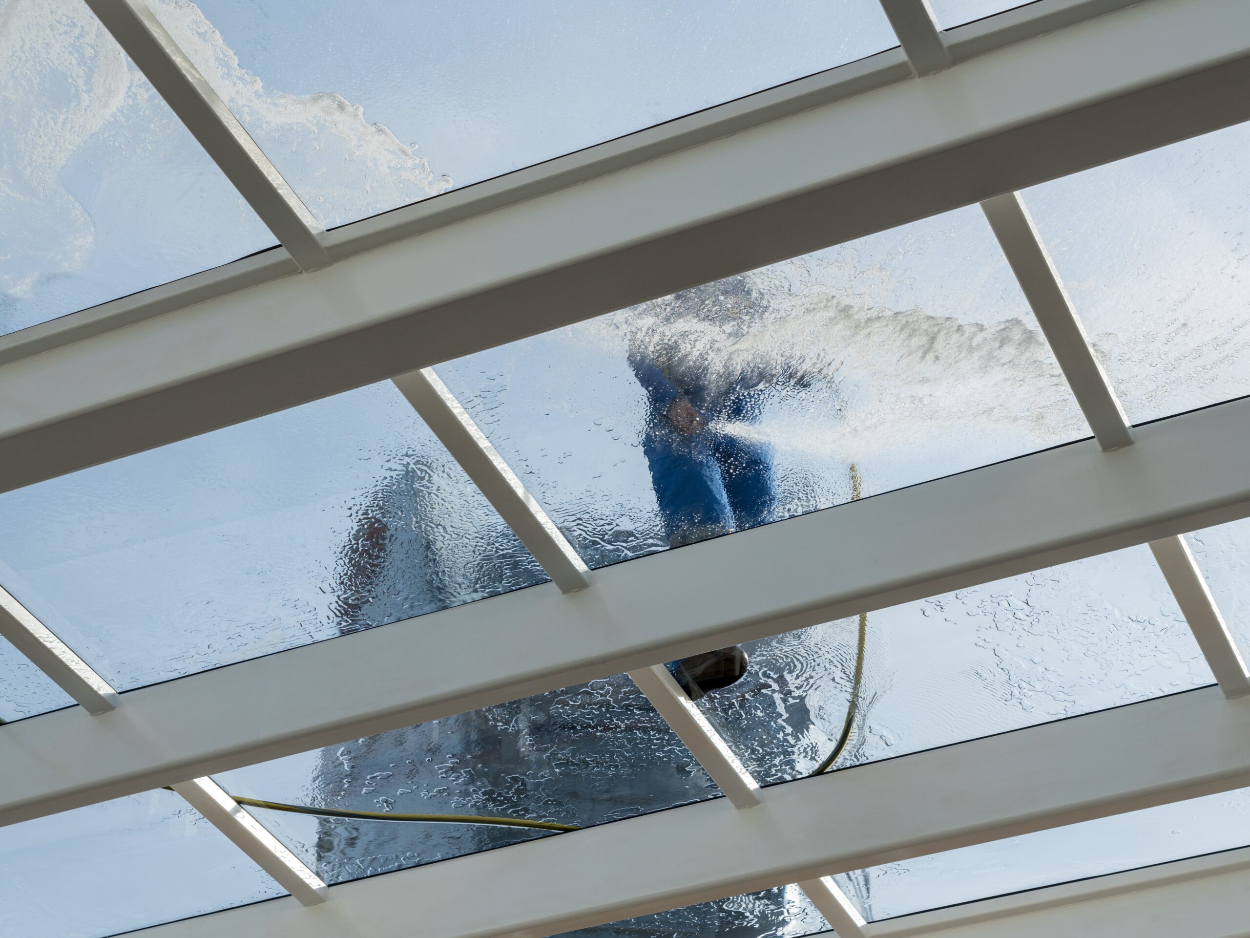 Male worker washing large expanse of glass roof over swimming pool with hose pipe